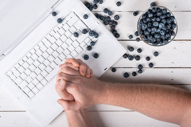 руки за компьютером и летняя голубая черника на деревянном белом столе. концепция здоровой закуски в offic - Фото, изображение