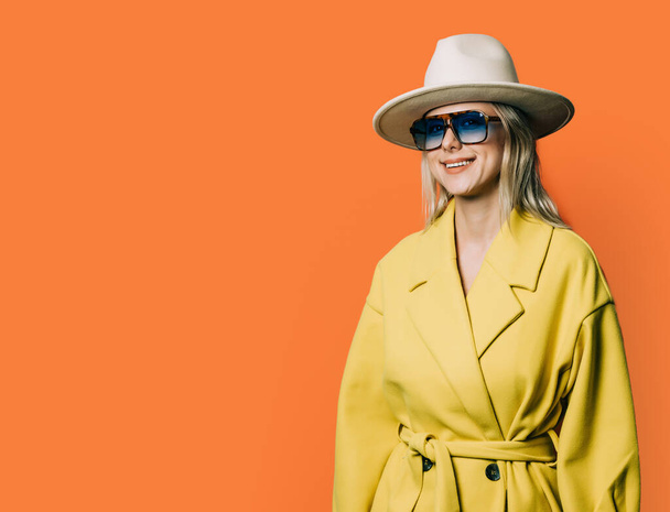 Stylish 80s woman in eyeglasses and hat with jacket on orange background - 写真・画像