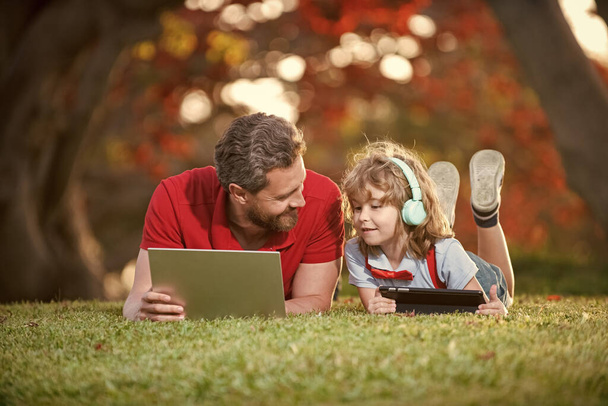 onnellinen perhe isä ja poika käyttää kannettavaa tietokonetta videopuhelun tai oppitunnin kuunnella musiikkia kuulokkeet puistossa, koulutus verkossa. - Valokuva, kuva