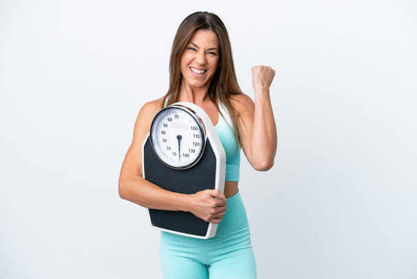 Middle age caucasian woman isolated on white background with weighing machine and doing victory gesture - 写真・画像