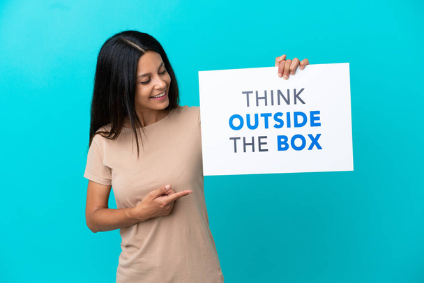 Молода жінка над ізольованим тлом тримає плакат з текстом Подумайте поза коробкою і вказуючи на нього
 - Фото, зображення