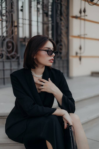 a brunette girl in a black jacket and dress and black glasses sits in the city with a handbag - Photo, image