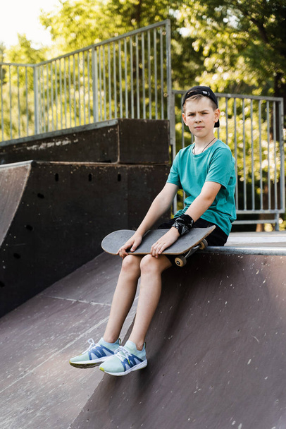 Criança com prancha de skate. Rapaz com skate sentado na rampa desportiva no parque de skate. Estilo de vida extremo. Anúncio criativo para lojas ou lojas de skate ou penny board - Foto, Imagem