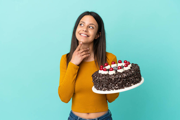 Young caucasian woman holding birthday cake isolated on blue background looking up while smiling - Zdjęcie, obraz