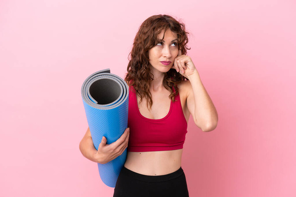 Young sport woman going to yoga classes while holding a mat isolated on pink background having doubts and thinking - Photo, image