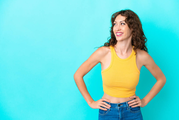 Young woman with curly hair isolated on blue background posing with arms at hip and smiling - Foto, Bild