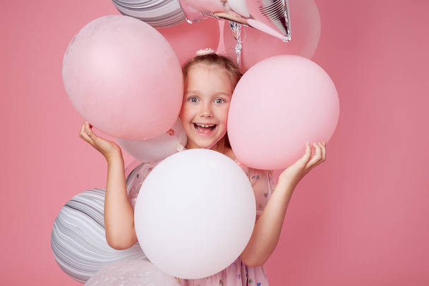 Joyful child girl in elegant tulle dress near the balloons. Birthday present. Funny face celebrates birthday party on pink background	 - Photo, Image
