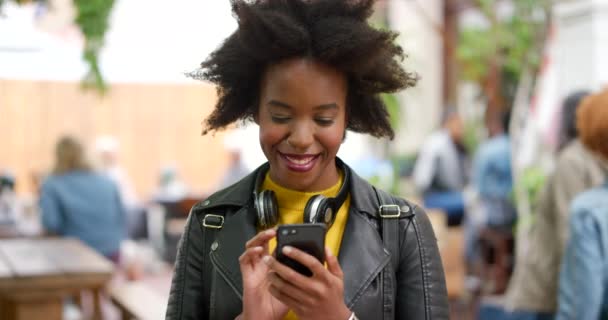 Jeune femme heureuse, branchée et élégante utilisant un téléphone pour envoyer un texte en ligne dans la ville. Une femme noire joyeuse, agitée et insouciante avec un afro souriant et naviguant sur le web tout en se tenant debout au centre-ville. - Séquence, vidéo