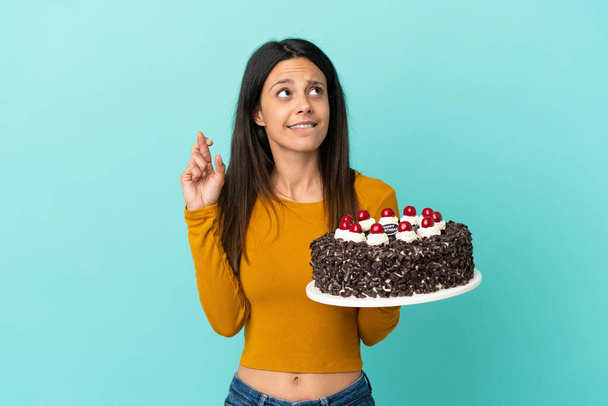 Young caucasian woman holding birthday cake isolated on blue background with fingers crossing and wishing the best - Foto, immagini
