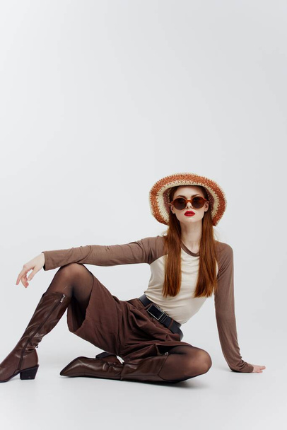 Un jeune modèle féminin mignon dans un chapeau bottes de lunettes est assis sur le sol dans un studio blanc. Photographie catalogue modèle. Photo de haute qualité - Photo, image