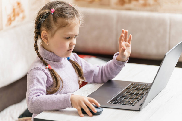 A beautiful cute little girl sits and communicates through a laptop with an interlocutor, telling something and waving her arms. - Photo, image