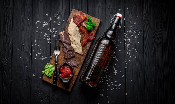Dried meat, jerky, with spicy sauce and a bottle of beer on a dark background. Beer snacks - Фото, изображение