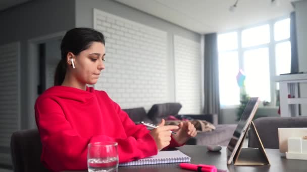 Beautiful caucasian female student is studying in college remotely. She is sitting at a table at home with a laptop and a notepad and concentrated is watching a video conference lesson - Felvétel, videó