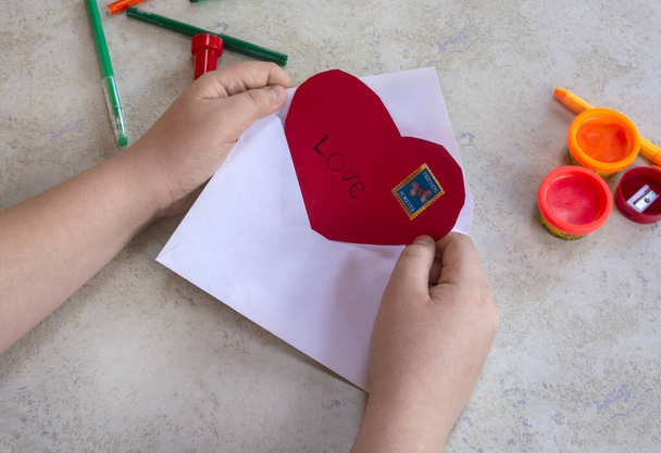 Valentijnsdag thema. Ambachtelijke concept. Kind stopt rood hart in envelop, maakt zich klaar voor Valentijn. Hoge kwaliteit foto - Foto, afbeelding