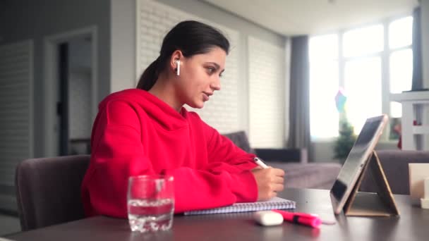 Beautiful caucasian female student is studying in college remotely. She is sitting at a table at home with a laptop and a notepad and concentrated is watching a video conference lesson - Video, Çekim