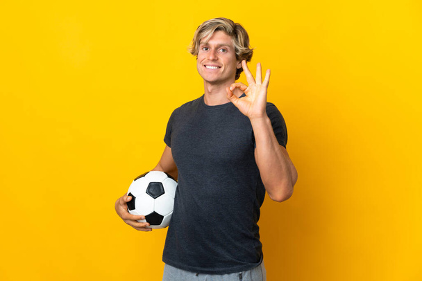 Hombre inglés sobre fondo amarillo aislado con pelota de fútbol y haciendo signo OK - Foto, imagen