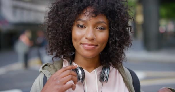 Portret van een trendy jonge zwarte vrouw met een zorgeloze en koele houding die buiten in de stad staat. Mooi, zelfverzekerd en vrolijk Afrikaans student glimlachen met krullend afro haar en koptelefoon. - Video