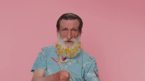 Mime senior man with flowers in gray-haired beard pulling an imaginary unreal invisible rope, puts lot of effort into showing how enduring, strong he is. Elderly grandfather on pink studio background - Filmagem, Vídeo