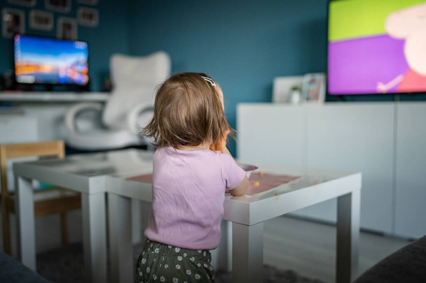 Vista posterior de una niña caucásica desconocida niño pequeño de pie en casa viendo la televisión mientras come espacio de copia infancia creciendo desarrollo concepto de ocio - Foto, imagen