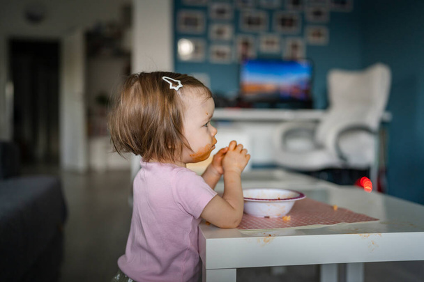 one girl small caucasian toddler female child daughter eating alone at the table at home while watching tv making mess on her face dirty sauce childhood growing up development concept copy space - Fotografie, Obrázek