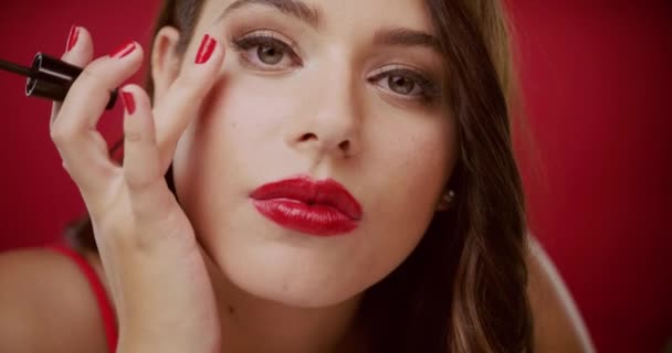 4k video footage of a beautiful young woman applying mascara against a red studio background. - Imágenes, Vídeo