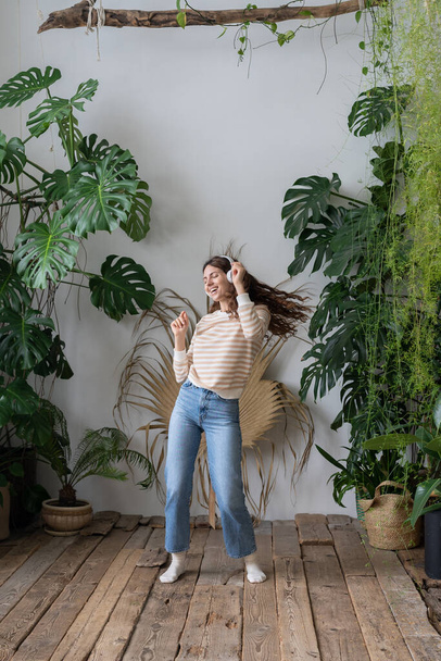 Positive young italian woman in wireless headphones listening to music with pleasure, enjoying the moment, relaxing, dancing on wooden floor in cozy home garden with monstera and tropical plants.  - Фото, изображение