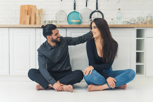Closeness and tenderness. Barefoot heterosexual couple - bearded formally dressed handsome Cuban man and long-haired caucasian casually dressed woman - sitting on the white kitchen floor. Man touching - Fotografie, Obrázek