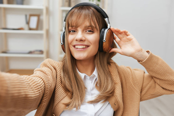 Cheerful young blonde student lady freelancer in warm sweater in headphones listen fav songs sitting in armchair at modern home interior. Music time Relaxing Cool playlist Concept. Wide selfie view - Photo, Image