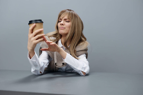 Несчастная молодая блондинка-студентка-фрилансер в тёплом свитере смотрит на кофе на рабочем месте в сером современном домашнем офисе. Кофе-брейк Lover Concept. Принято. Холодное предложение - Фото, изображение