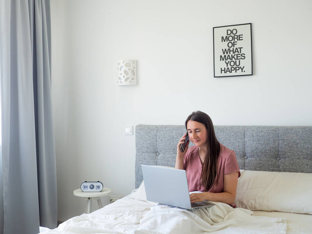 Женщина, работающая с ноутбуком на кровати. Концепция цифровой рекламы и работы из дома. Молодая деловая женщина, работающая за компьютером, лежит в красивой белой и солнечной спальне. вид с пространством для копирования - Фото, изображение