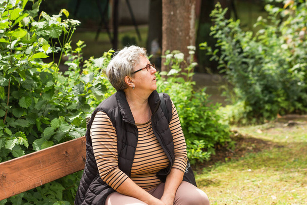Πλευρική άποψη της προνοητικής ηλικιωμένης γυναίκας με γυαλιά που κάθεται και ξεκουράζεται στον πάγκο του πάρκου. Γκρι μαλλιά γυναίκα σε casual ρούχα ξοδεύει τον ελεύθερο χρόνο της στο πράσινο καλοκαιρινό πάρκο. Έννοια της τρίτης ηλικίας. - Φωτογραφία, εικόνα