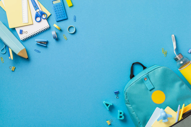 Top view photo of backpack notebooks pencil-case scissors drink bottle plastic alphabet letters binder clips plane shaped sharpener pens adhesive tape stapler ruler calculator isolated blue background - Photo, Image
