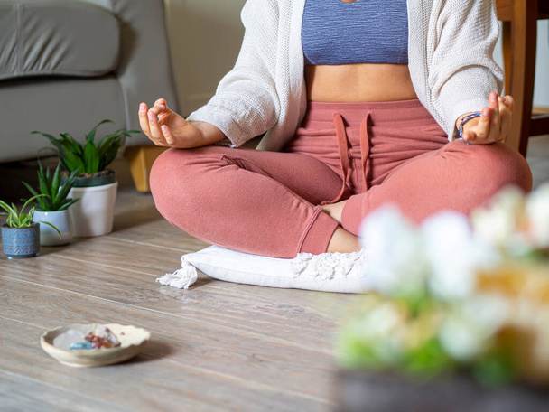 Woman practicing meditation at home with crystals and plants. Unrecognizable young adult meditating in quiet and relaxing room sitting in lotus position on a pillow. Mental health and stress relief. - Photo, Image
