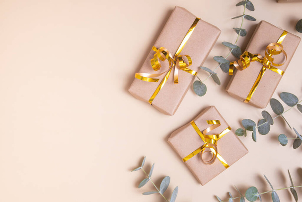 Geschenkverpackungen in Kraftpapier mit goldener Schleife und Eukalyptus auf pastellbeigem Hintergrund. Feriengeschenkkonzept. Ansicht von oben. - Foto, Bild