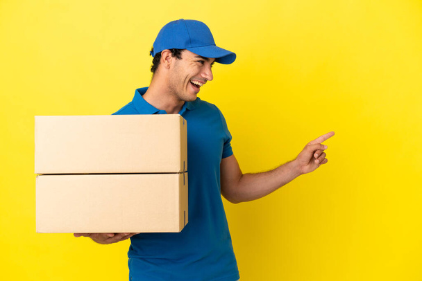 Delivery man over isolated yellow wall pointing finger to the side and presenting a product - Photo, Image