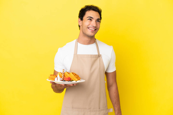 El camarero del restaurante sostiene gofres sobre un fondo amarillo aislado pensando en una idea mientras mira hacia arriba - Foto, Imagen