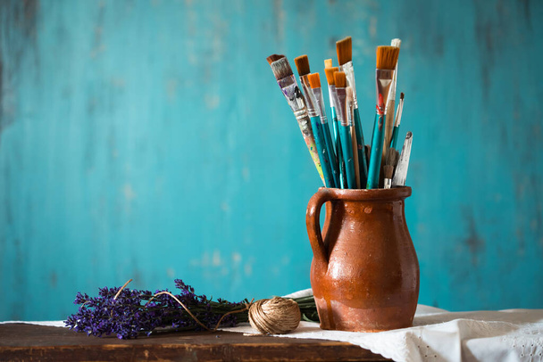 Still life of paint brushes  in clay jug an lavender flower on vintage turquoise background - Photo, Image