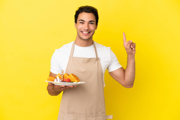 Restaurant waiter holding waffles over isolated yellow background pointing up a great idea - Foto, afbeelding
