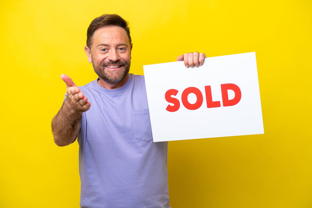 Middle age caucasian man isolated on yellow background holding a placard with text SOLD making a deal - Foto, Imagen