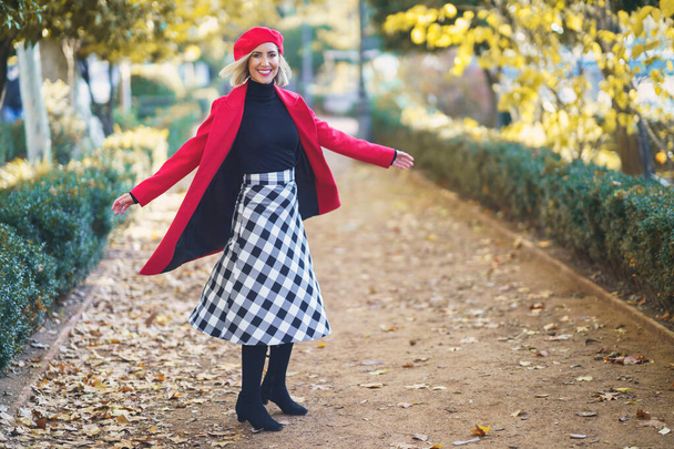 Full body of positive female in red outfit looking at camera while spinning around on pathway in autumn park with trees - Photo, image