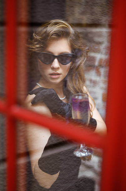 A beautiful Caucasian girl in sunglasses is drinking a cocktail near the bar. Taken through the glass of the door - Foto, Imagem