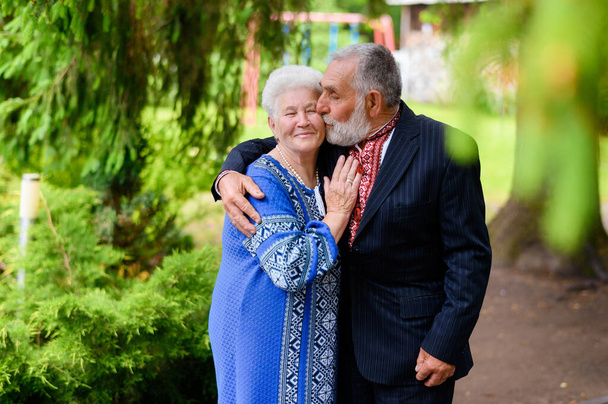 An old gray-haired woman hugs an old gray-haired man and sweetly kisses on the background of a park in Ukrainian traditional clothes - Photo, image