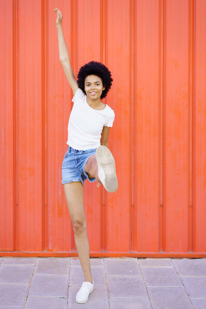 Cuerpo completo de alegre mujer afroamericana con peinado afro en ropa casual mirando a la cámara con la pierna levantada sobre fondo rojo - Foto, Imagen
