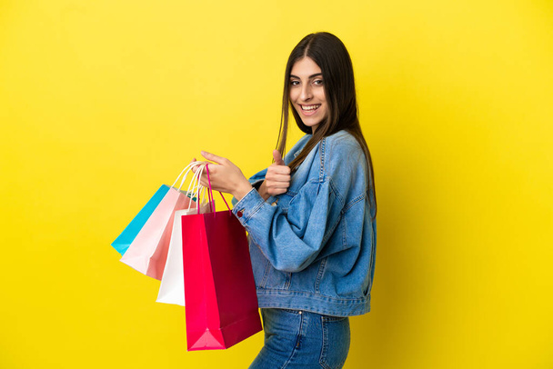 Young caucasian woman isolated on blue background holding shopping bags and with thumb up - 写真・画像