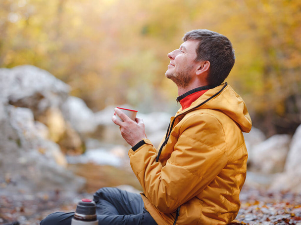 Sonbaharda seyahat ve yolculuk konsepti. Macera ve aktif yaşam tarzı. Turist yürüyüşü. Sarı ceketli beyaz adam ormanda nehir kenarında oturmuş termostan çay içiyor. - Fotoğraf, Görsel