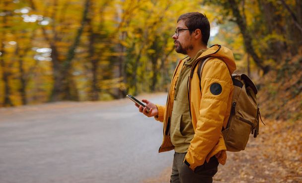 Young handsome man in autumn forest with smartphone. young hipster guy with backpack , traveller standing in woods, Hiking, Forest, Journey, active healthy lifestyle, adventure, vacation concept. - Φωτογραφία, εικόνα