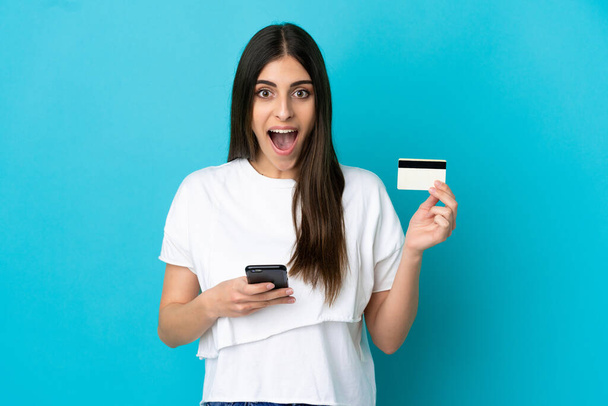 Mladá běloška izolované na modrém pozadí nákup s mobilem a drží kreditní kartu s překvapeným výrazem - Fotografie, Obrázek