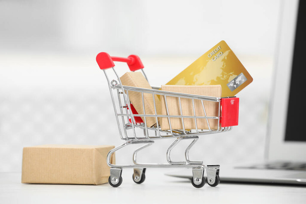 Online payment concept. Small shopping cart with bank card, boxes and laptop on white table, closeup - Foto, Bild