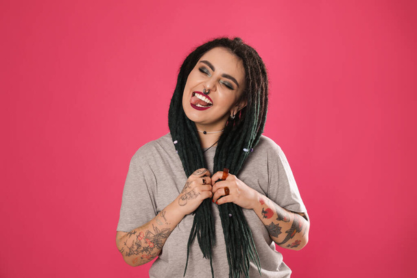 Beautiful young woman with tattoos on arms, nose piercing and dreadlocks against pink background - Valokuva, kuva
