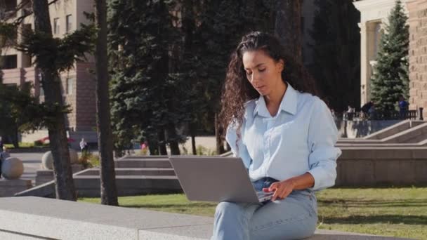 Σοβαρή νεαρή ελκυστική γυναίκα ελεύθερος επαγγελματίας εργαζόμενος κάθεται βεράντα στην πόλη κτίριο φόντο πληκτρολογώντας email κάνει εργασίες γραφείου χρησιμοποιώντας φορητό υπολογιστή app e-learning μακρινή εργασία σε απευθείας σύνδεση chat στο δρόμο - Πλάνα, βίντεο